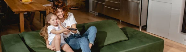 Panoramic orientation of cute kid holding remote controller and sitting near redhead mother on sofa — Stock Photo