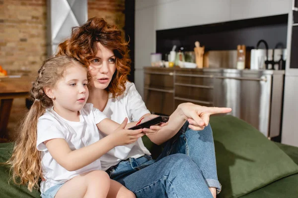 Cute kid holding remote controller and sitting near redhead mother pointing with finger in living room — Stock Photo