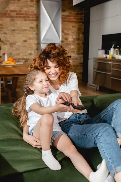 Cute kid holding remote controller and sitting near cheerful mother on sofa — Stock Photo