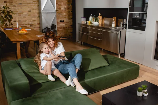 Niño feliz sosteniendo mando a distancia y sentado cerca de la madre alegre en el sofá - foto de stock