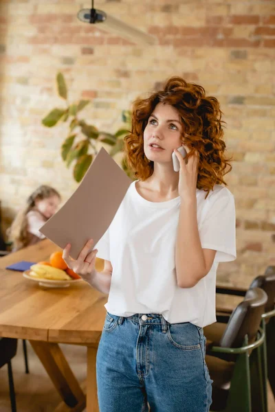 Selective focus of redhead woman talking on smartphone and holding folder near daughter at home — Stock Photo