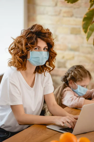 Selective focus of redhead freelancer mother in medical mask using laptop near daughter — Stock Photo