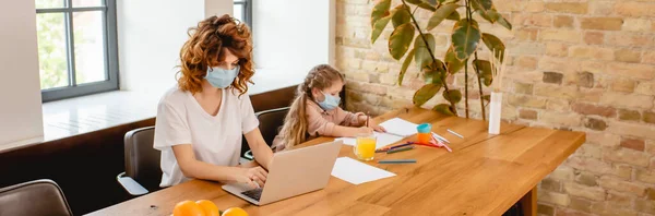 Tiro panorâmico de mãe freelancer encaracolado em máscara médica usando laptop perto filha desenho em casa — Fotografia de Stock