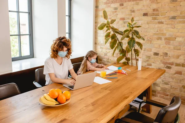 Curly freelancer mother in medical mask using laptop near daughter drawing at home — Stock Photo