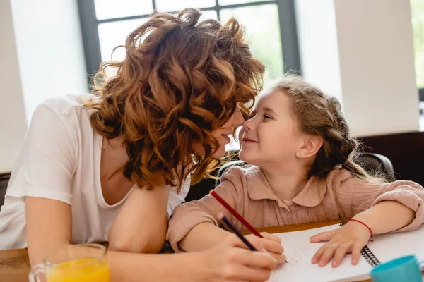 Messa a fuoco selettiva di madre riccia toccare nasi e disegno con figlia a casa — Foto stock