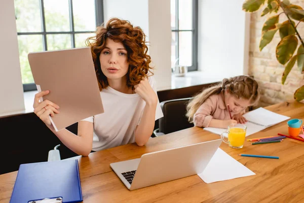 Rizado freelancer madre celebración carpeta cerca de portátil e hija dibujo en casa - foto de stock