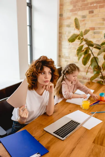 Freelancer encaracolado mãe segurando pasta perto laptop e criança desenho em casa — Fotografia de Stock