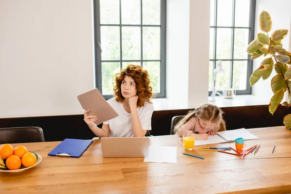 Ruiva freelancer mãe segurando pasta perto laptop e criança desenho em casa — Fotografia de Stock