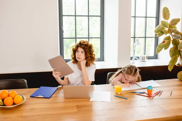 Pelirroja freelancer madre celebración carpeta cerca de la computadora portátil y el niño dibujo en casa - foto de stock