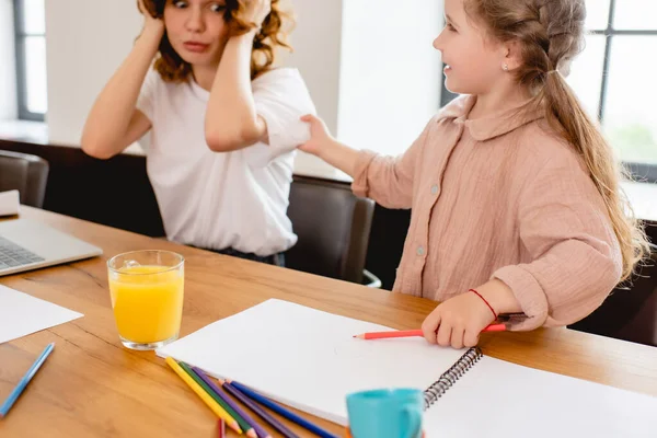 Enfoque selectivo de la madre pelirroja cubriendo oídos cerca de la computadora portátil y la hija en casa - foto de stock