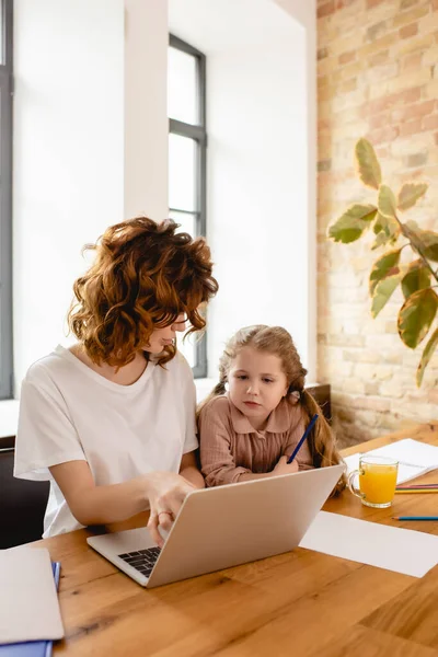 Nette Tochter schaut auf Laptop, während Mutter von zu Hause aus arbeitet — Stockfoto
