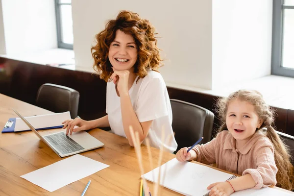 Enfoque selectivo de feliz y rizado freelancer madre sonriendo cerca de niño dibujo en casa - foto de stock