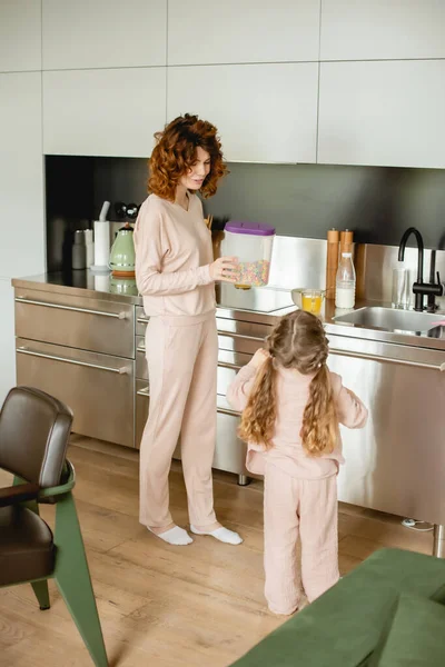 Vue arrière de l'enfant près de la mère bouclée contenant avec de savoureux flocons de maïs dans la cuisine — Photo de stock