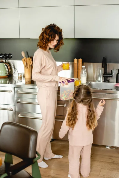 Back view of kid near curly mother holding container with tasty corn flakes and glass of orange juice in kitchen — Stock Photo