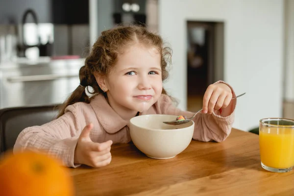 Foco seletivo de criança segurando colher perto da tigela com flocos de milho e vidro de suco de laranja — Fotografia de Stock