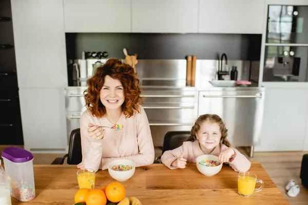 Happy mother and cute daughter holding spoons near bowls with corn flakes and glasses of orange juice — Stock Photo