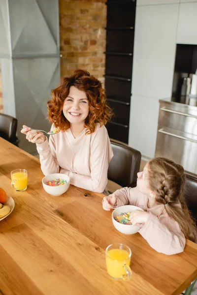 Cheerful mother and cute daughter holding spoons near bowls with corn flakes and glasses of orange juice — Stock Photo