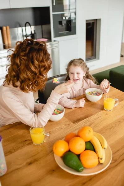 Mãe encaracolado e feliz filha segurando colheres perto de tigelas com flocos de milho e copos de suco de laranja — Fotografia de Stock