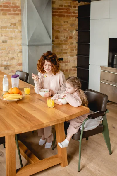 Madre rizada y linda hija sosteniendo cucharas cerca de cuencos con copos de maíz, frutas y vasos de jugo de naranja - foto de stock