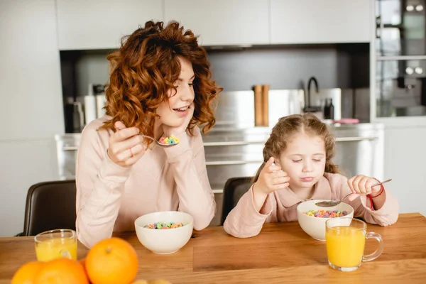 Curly mother and adorable kid holding spoons near bowls with corn flakes and glasses of orange juice — Stock Photo