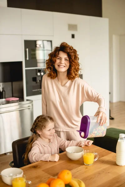 Cheerful mother holding container with corn flakes near daughter, bottle with milk and glasses of orange juice — Stock Photo