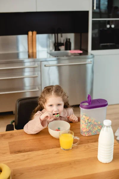 Lindo niño comiendo copos de maíz cerca de vaso de jugo de naranja y botella con leche - foto de stock