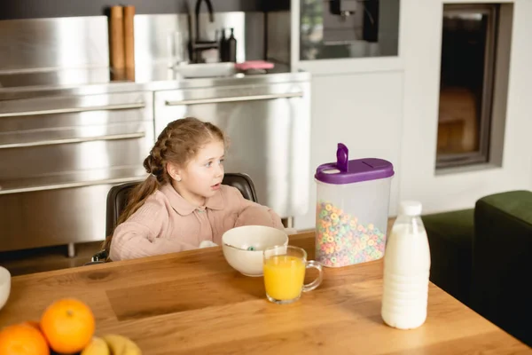 Glückliches Kind schaut weg in der Nähe von Cornflakes, einem Glas Orangensaft und einer Flasche mit Milch — Stockfoto