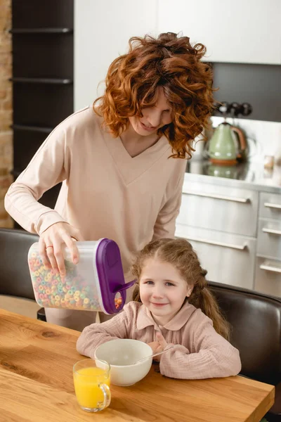 Happy mother holding container with delicious corn flakes near bowl, glass and cheerful daughter — Stock Photo