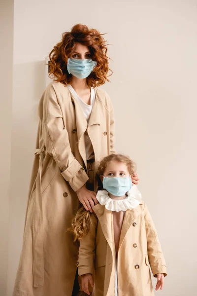 Curly mother and daughter in medical masks standing in trench coats — Stock Photo