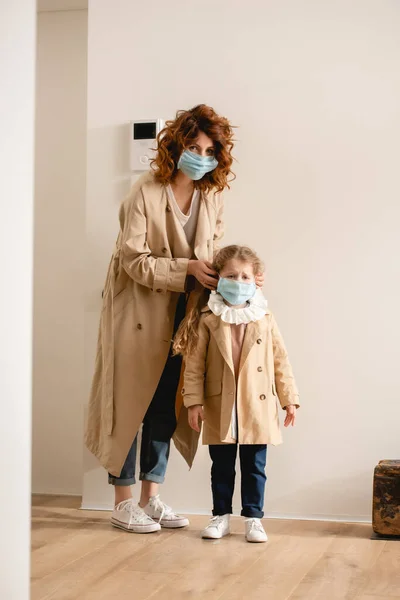 Curly mother in trench coat wearing medical mask on cute daughter — Stock Photo