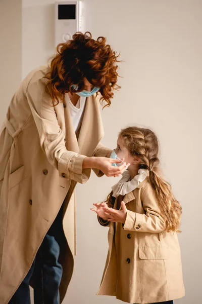 Mãe encaracolado em casaco de trincheira vestindo máscara médica na filha adorável — Fotografia de Stock