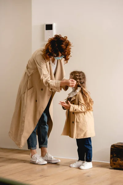 Curly mother in trench coat wearing medical mask on kid — Stock Photo