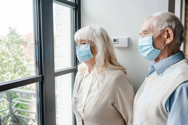 Sad elderly couple in medical masks looking through window during self isolation — Stock Photo