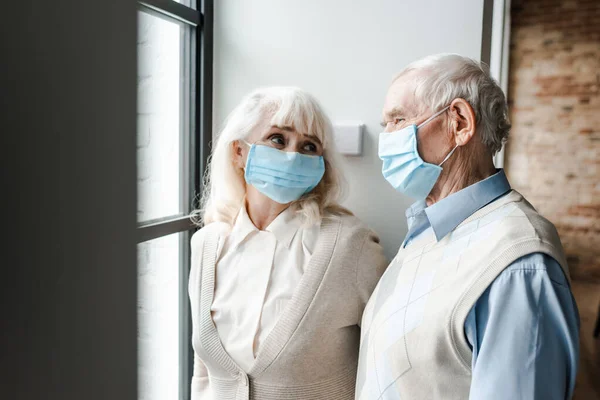 Elderly couple in medical masks looking through window during quarantine — Stock Photo