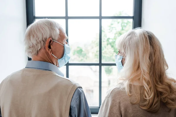 Вид ззаду сумної пари літніх людей в медичних масках, які дивляться через вікно під час самоізоляції — стокове фото