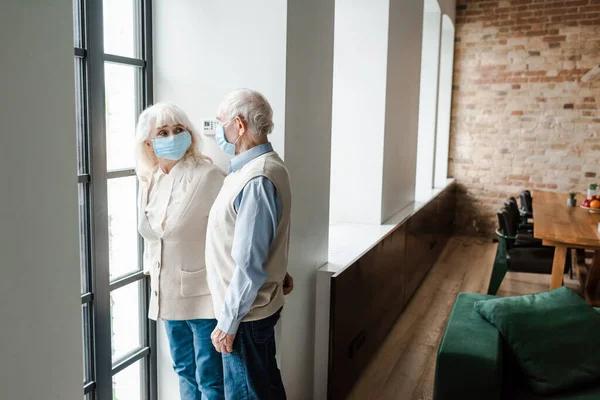 Upset senior couple in medical masks standing near through window during quarantine — Stock Photo