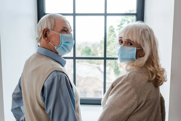 Sad senior couple in medical masks standing near window during self isolation — Stock Photo