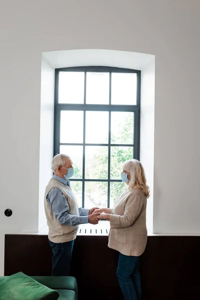 Sad elderly couple in medical masks holding hands and standing near window during quarantine — Stock Photo
