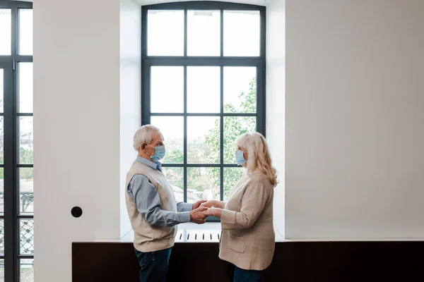 Casal sênior em máscaras médicas de mãos dadas e de pé perto da janela durante o auto-isolamento — Fotografia de Stock