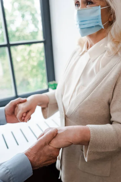 Ausgeschnittene Ansicht eines älteren Ehepaares in medizinischen Masken, das sich während der Selbstisolierung zu Hause an den Händen hält — Stockfoto