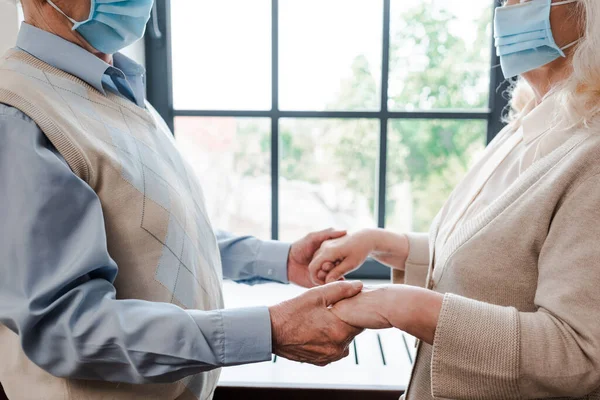 Ausgeschnittene Ansicht eines älteren Ehepaares in medizinischen Masken, das sich während der Selbstisolierung zu Hause an den Händen hält — Stockfoto