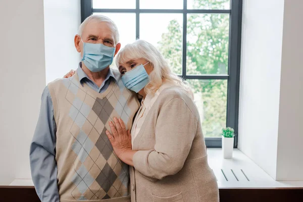 Upset elderly couple in medical masks hugging at home during self isolation — Stock Photo