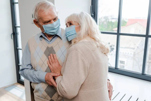 Triste pareja de ancianos en máscaras médicas abrazándose en casa durante el auto aislamiento - foto de stock