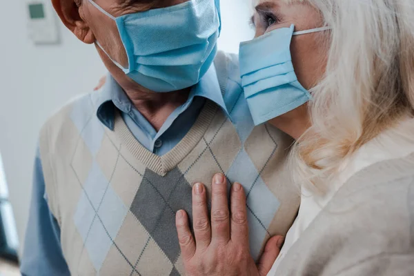 Обрезанный вид пожилой пары в медицинских масках, обнимающейся дома во время самоизоляции — стоковое фото