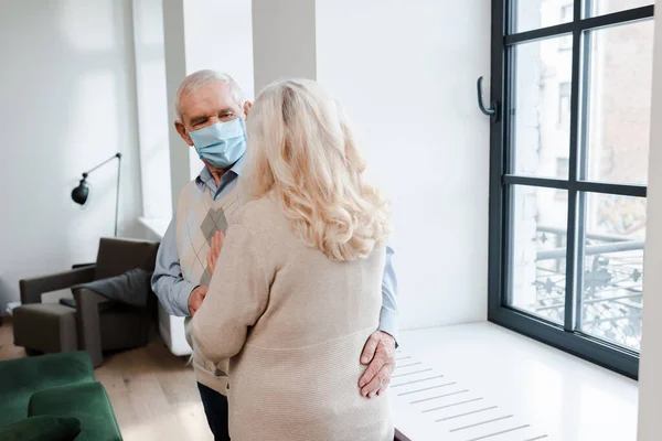 Senior couple in medical masks hugging at home during self isolation — Stock Photo