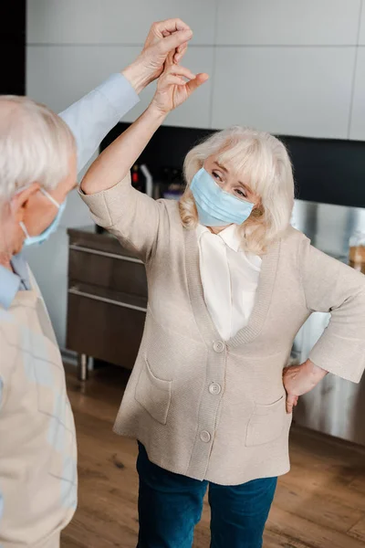 Happy elderly couple in medical masks dancing at home during self isolation — Stock Photo