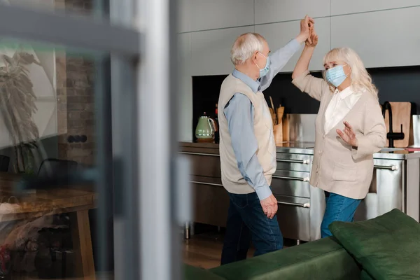 Pareja de ancianos en máscaras médicas bailando en la cocina durante el autoaislamiento - foto de stock