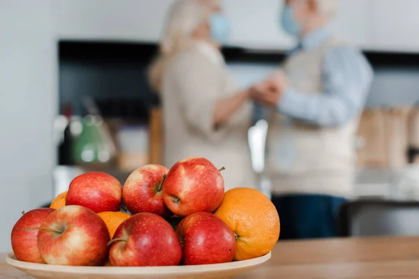 Seniorpaar in medizinischen Masken tanzt in der Küche mit Früchten während der Selbstisolierung, selektiver Fokus — Stockfoto