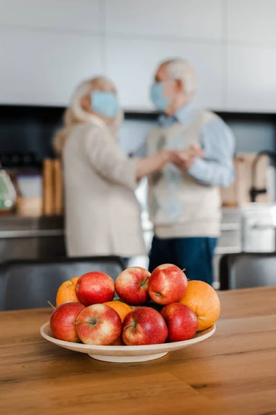 Selective focus of elderly couple in medical masks dancing on kitchen with fruits during quarantine — Stock Photo