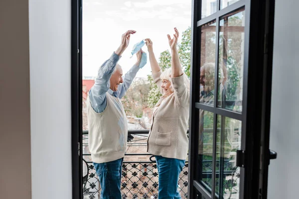 Excitada pareja de ancianos sosteniendo máscaras médicas mientras está de pie en el balcón después de auto aislamiento - foto de stock